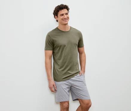 Mann mit lockigem Haar, olivgrünem T-Shirt und grauen Shorts steht vor einer weißen Wand und lächelt.