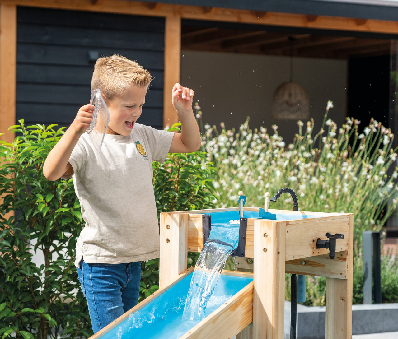 Junge spielt mit einem hölzernen Wasserspieltisch im Freien, Wasser spritzt.