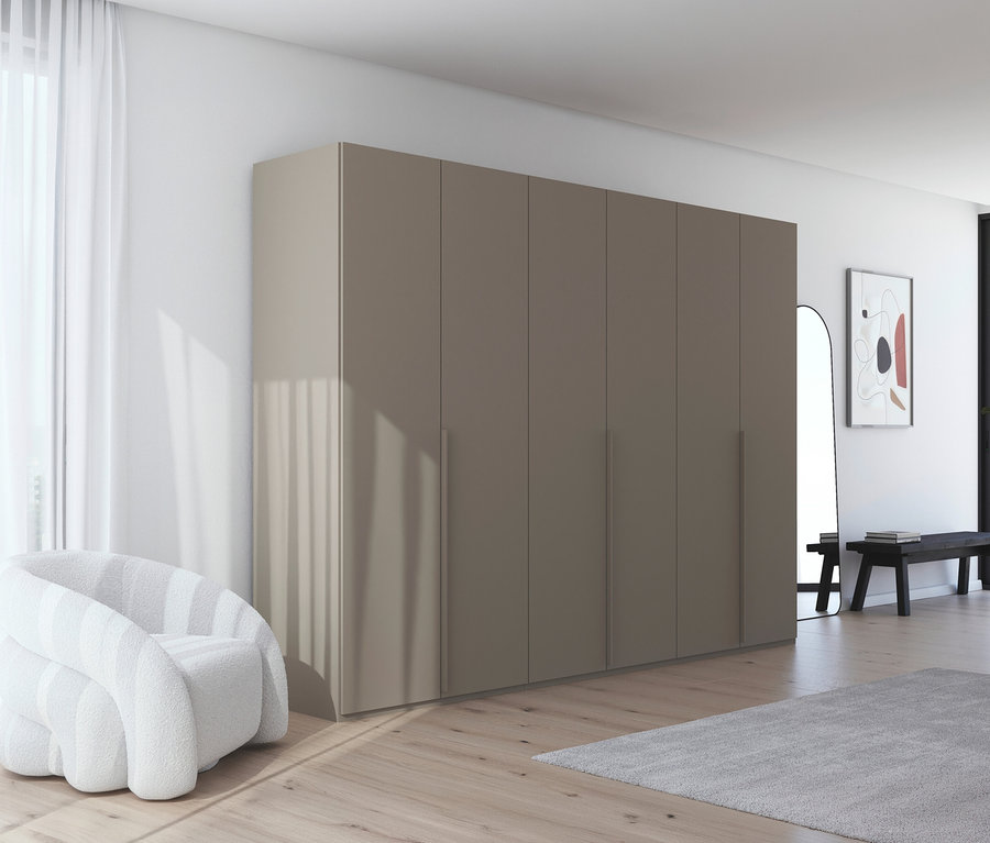 Beiger Kleiderschrank mit mehreren Türen neben weißem Sessel und Vorhang. Im Hintergrund sind ein Spiegel und eine Bank zu sehen.