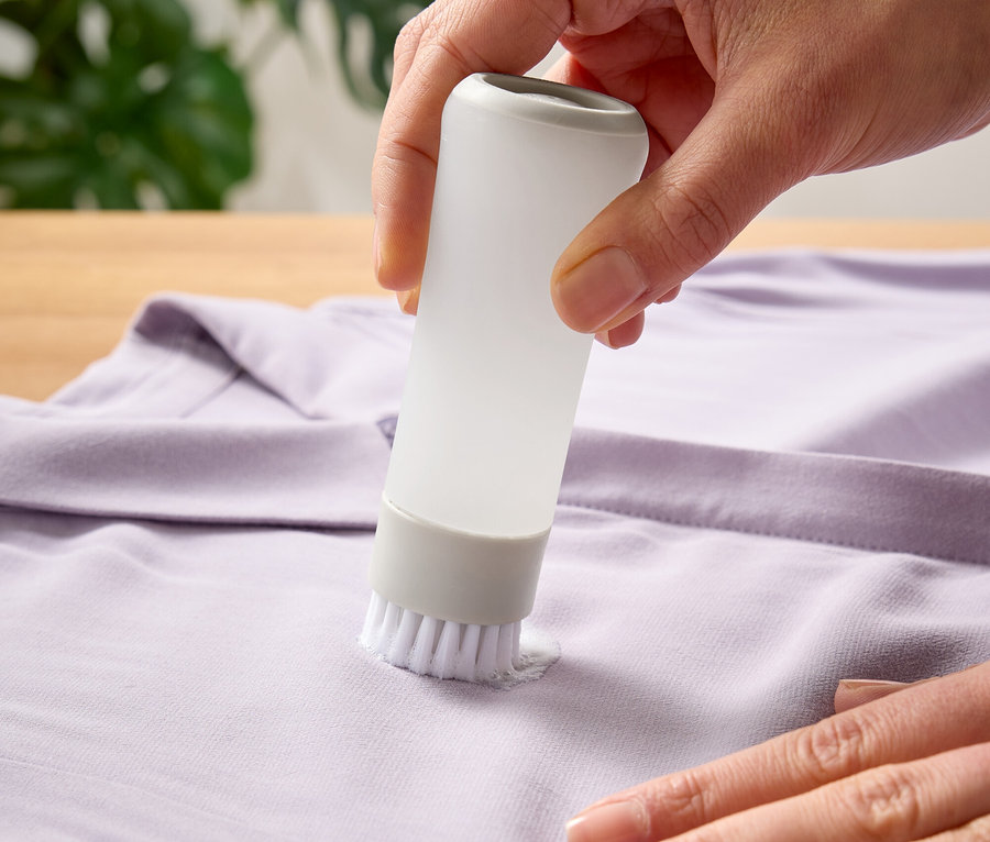 Eine Hand verwendet eine Bürste mit Behälter, um einen Fleck auf einem lila T-Shirt zu bearbeiten.
