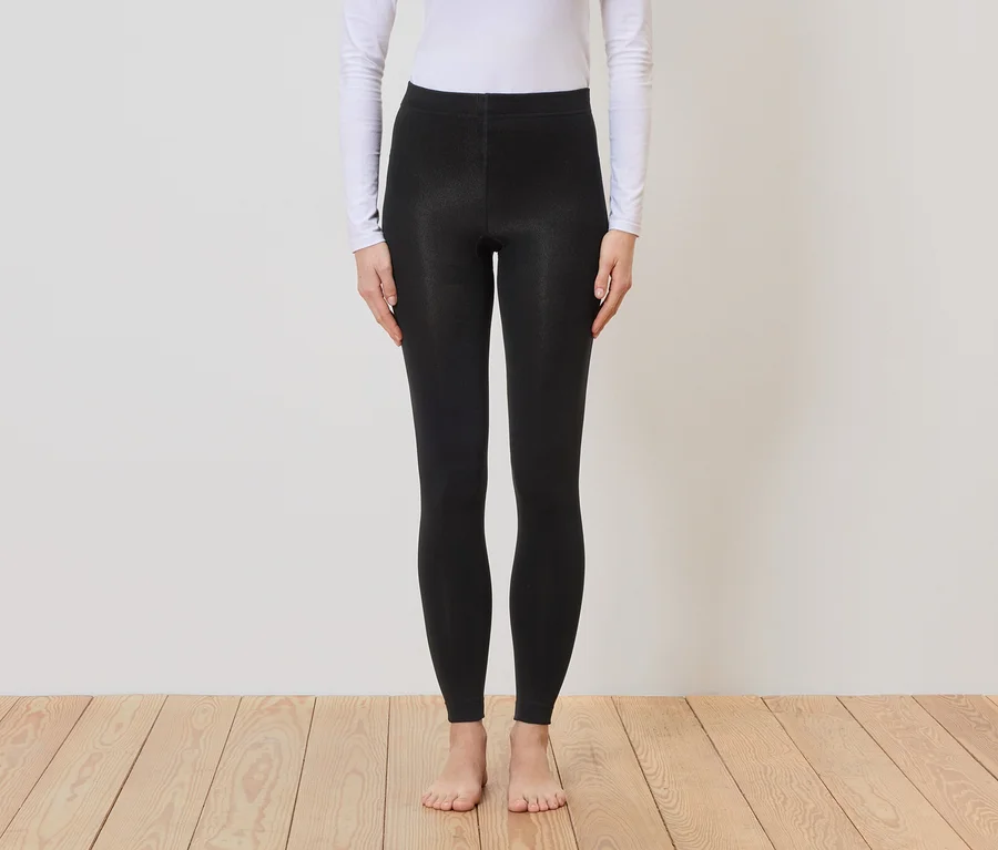 Eine barfüßige Person mit weißem, langärmligem Oberteil und schwarzer Leggings steht vor einer Wand und einem Holzboden.