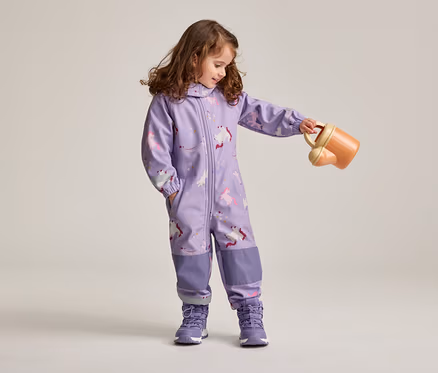 Mädchen mit braunen, lockigen Haaren in lila Overall mit Einhornmuster und lila Schuhen hält eine orangefarbene Gießkanne.