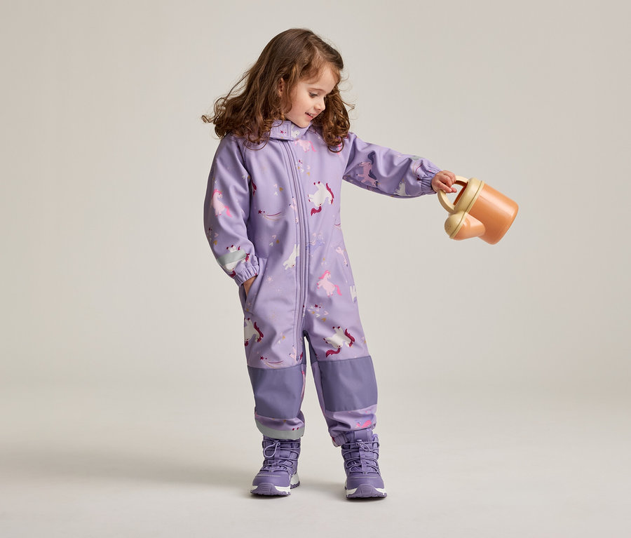 Mädchen mit braunen, lockigen Haaren in lila Overall mit Einhornmuster und lila Schuhen hält eine orangefarbene Gießkanne.