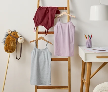 Eine Holzleiter lehnt an einer Wand, an der drei Tops hängen. Links steht ein Steckenpferd, rechts ein Schreibtisch mit Stiften und Notizbuch.