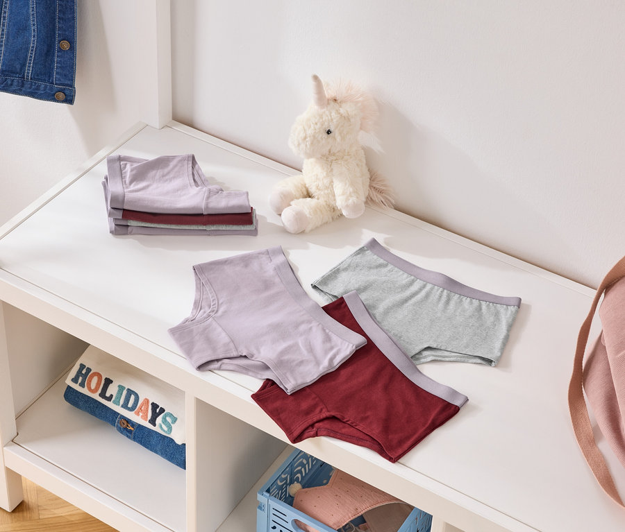 Auf einem Regal liegen gefaltete und ausgebreitete Unterhosen in Lila, Grau und Burgunderrot, neben einem Stoffeinhorn und einer rosafarbenen Tasche.