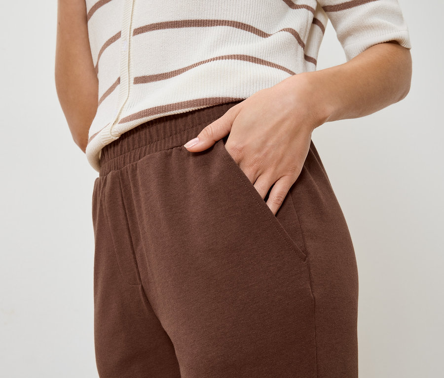 Nahaufnahme einer Person mit einer Hand in der Tasche einer braunen Jogginghose, dazu ein gestreiftes Oberteil.