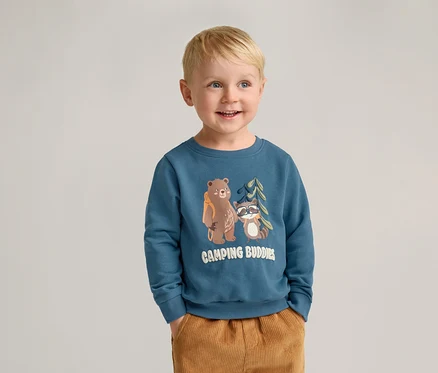 Lächelnder Junge mit blonden Haaren trägt ein blaues Sweatshirt mit Bär- und Waschbär-Motiv und braune Hosen.