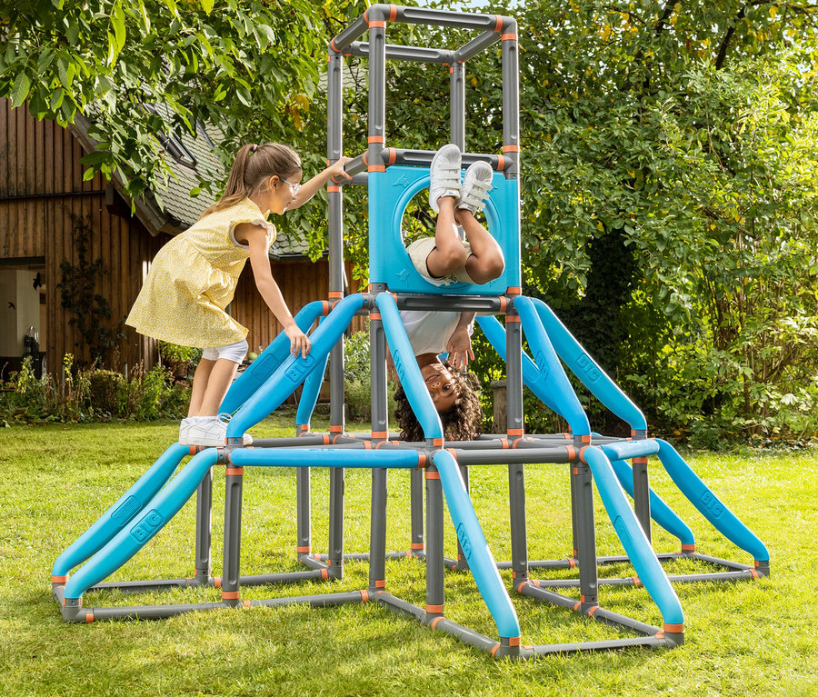 Zwei Kinder spielen auf einem grauen Klettergerüst mit blauen und orangen Akzenten im Garten.