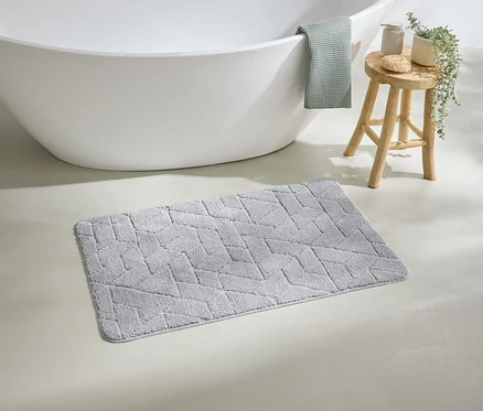 Ein grauer, geometrisch gemusterter Badteppich liegt vor einer weißen Badewanne. Ein grünes Handtuch hängt über dem Rand der Wanne.