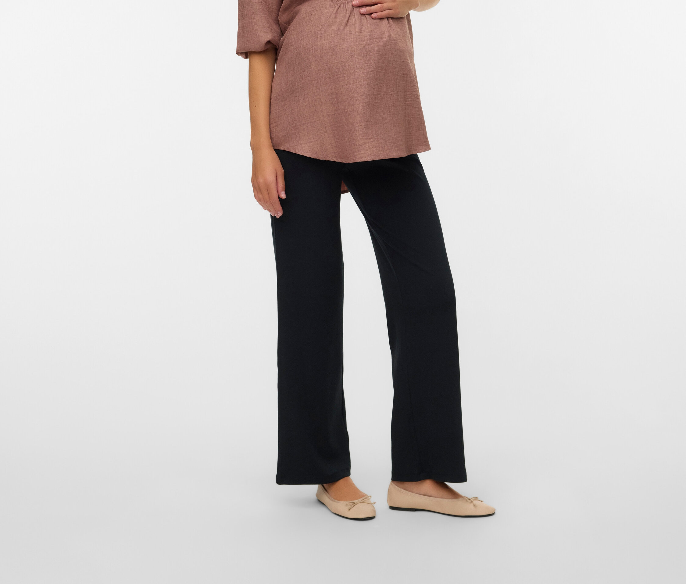 Schwangere Frau mit hellbrauner Bluse und schwarzen Hosen, die ihre Hände auf ihren Bauch legt.