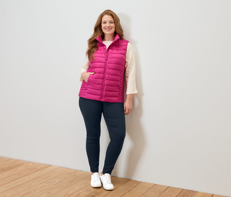 Frau in rosafarbener Steppweste, beiger Bluse, blauen Jeans und weißen Turnschuhen steht an einer weißen Wand.