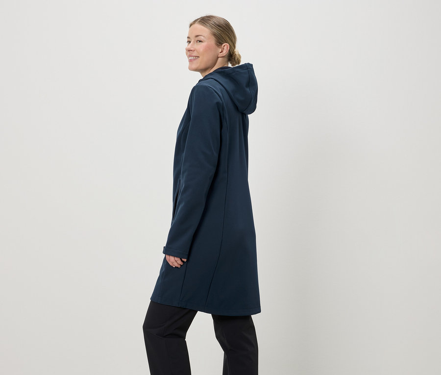 Eine Frau mit Pferdeschwanz trägt einen dunkelblauen Softshellmantel mit Kapuze und schwarze Hosen vor einem hellen Hintergrund.