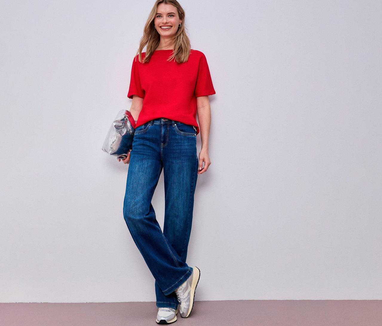 Frau mit rotem Street One Kurzarmshirt, blauen Jeans und silbernen Sneakern, eine silberne Tasche haltend.