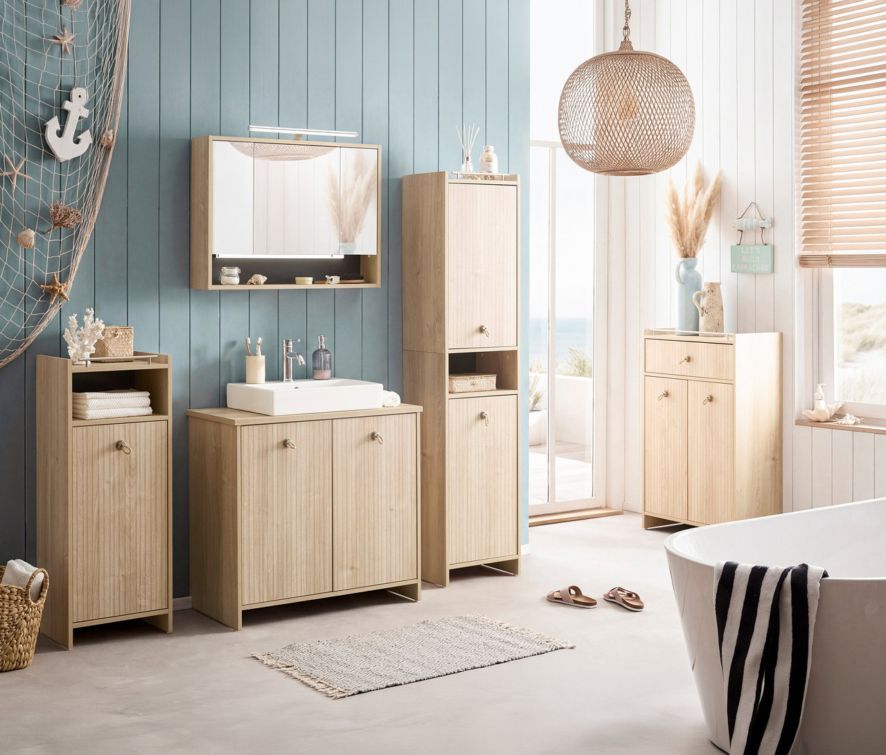 Badezimmer mit Waschtisch, Spiegelschrank, Hochschrank und Badewanne in hellem Holzdekor. Dekorationen wie Seestern und Anker sind angebracht.