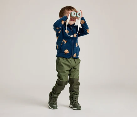 Ein Junge mit einer marineblauen Fleecejacke mit Bärenmuster schaut durch ein Spielzeugfernglas. Er trägt grüne Hosen und Stiefel.