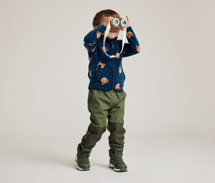 Ein Junge mit einer marineblauen Fleecejacke mit Bärenmuster schaut durch ein Spielzeugfernglas. Er trägt grüne Hosen und Stiefel.