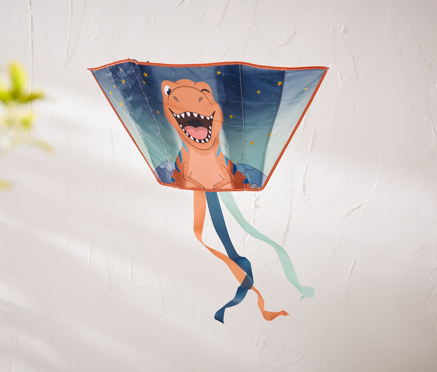 Ein trapezförmiger Drachen mit einem lachenden orangefarbenen Dinosaurier und Sternen hängt an einer strukturierten weißen Wand. Unten hängen orange, dunkelblaue und hellblaue Bänder.