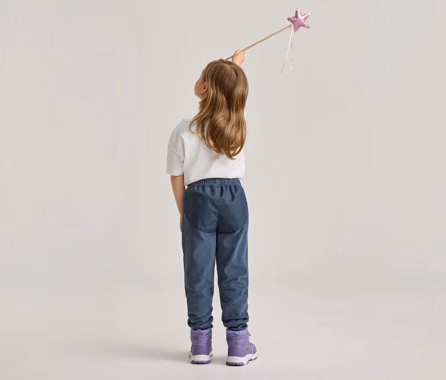 Rückansicht eines Mädchens mit blonden Haaren, das einen Zauberstab mit rosa Stern hält. Sie trägt blaue Hosen und lila Stiefel.