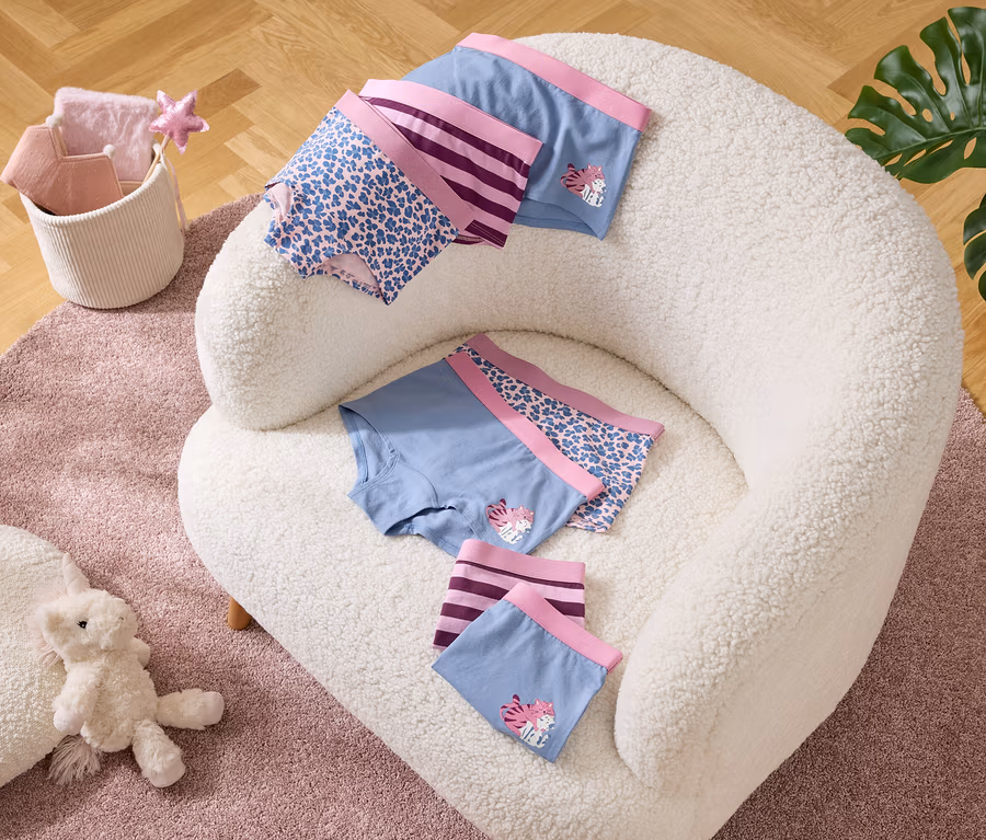 Auf einem Sessel liegen Unterhosen in Blau mit lila Bündchen und Mustern. Daneben ein Korb mit Textilien und ein Teddy auf einem Teppich.