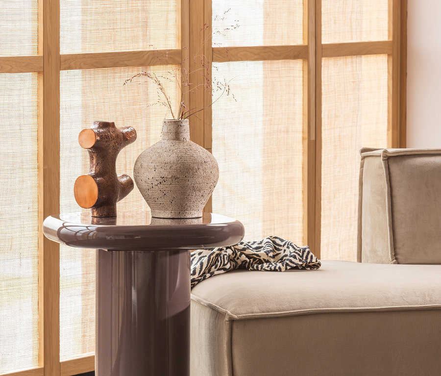Ein brauner Beistelltisch mit einer strukturierten hellen Vase und einer metallischen Skulptur. Rechts daneben steht ein beiges Sofa mit einem schwarz-weiß gemusterten Überwurf vor einem Fenster mit Holzrahmen und gewebten Jalousien.