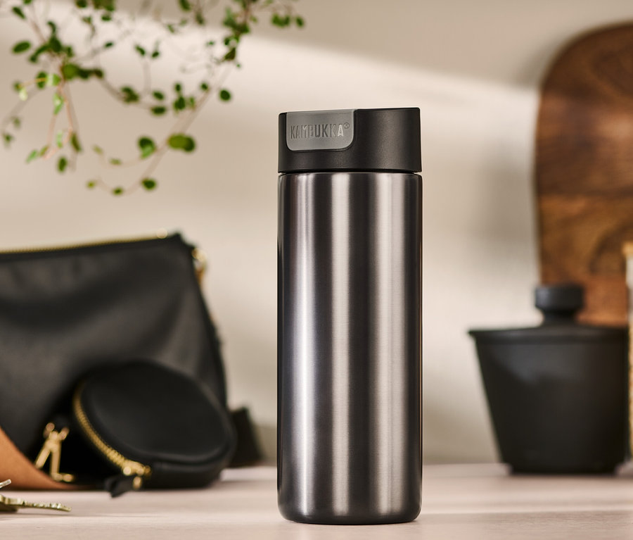Ein grauer Kambukka To Go Becher, 500 ml, steht auf einem Tisch neben einer Handtasche, einem Münzfach und Schlüsseln.