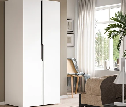 Weißer zweitüriger Kleiderschrank mit schwarzen Griffen in einem Raum mit Fenster, weißen Vorhängen, Holzstuhl, Bett und gestreiftem Teppich.