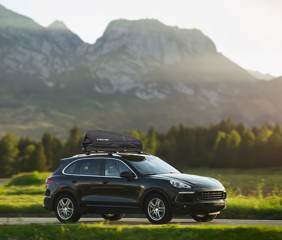 Schwarzer SUV fährt auf einer Straße mit einer schwarzen FISCHER Dachbox auf dem Dach, vor einer Kulisse aus sonnenbeschienenen Bergen und Wäldern.