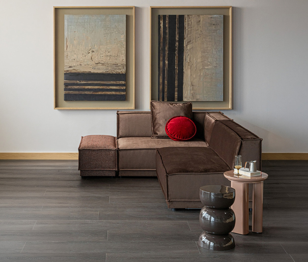 Ein modernes Wohnzimmer mit einem braunen L-förmigen Ecksofa, einem roten runden Kissen, zwei abstrakten Gemälden an der hellen Wand und zwei Beistelltischen. Auf einem Tisch stehen ein Glas und eine silberne Kanne.