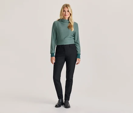 Eine Frau steht in einem grün-weiß gestreiften Kapuzenpullover, schwarzen Hosen und schwarzen Stiefeln.