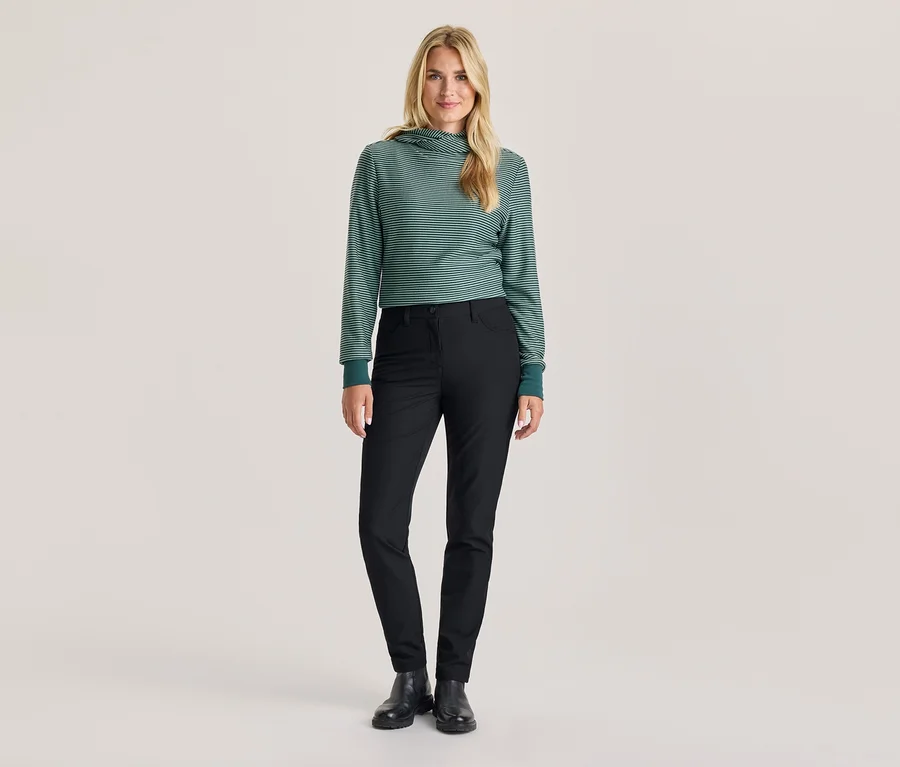 Eine Frau steht in einem grün-weiß gestreiften Kapuzenpullover, schwarzen Hosen und schwarzen Stiefeln.