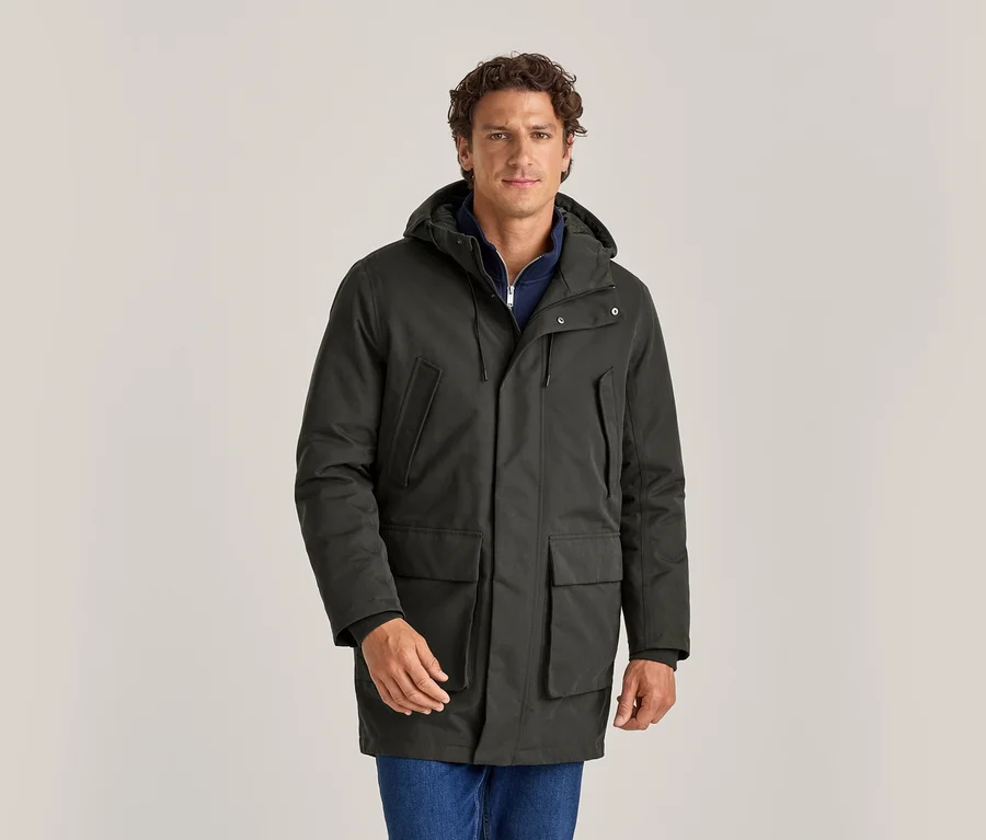 Mann trägt einen dunkelgrünen Kapuzenparka über einem blauen Oberteil und blauen Jeans, steht und blickt nach vorne.