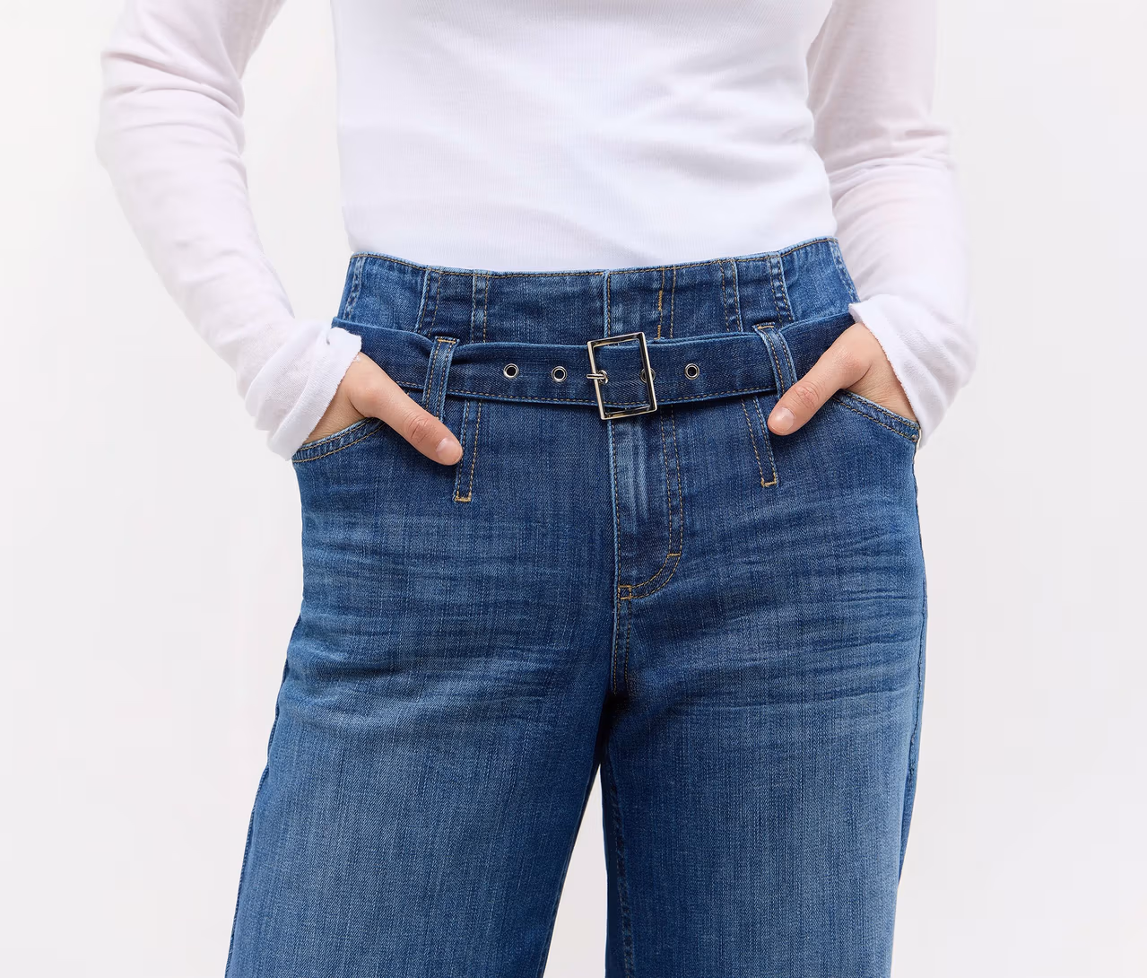 Person trägt blaue High-Waist-Jeans mit integriertem Gürtel und weißes Langarmshirt, Hände in den Hosentaschen.