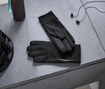 Schwarzer Fahrradhelm, dunkle Trinkflasche, schwarze Handschuhe mit reflektierenden Punkten und schwarze Kopfhörer liegen auf einer grauen Betonoberfläche.