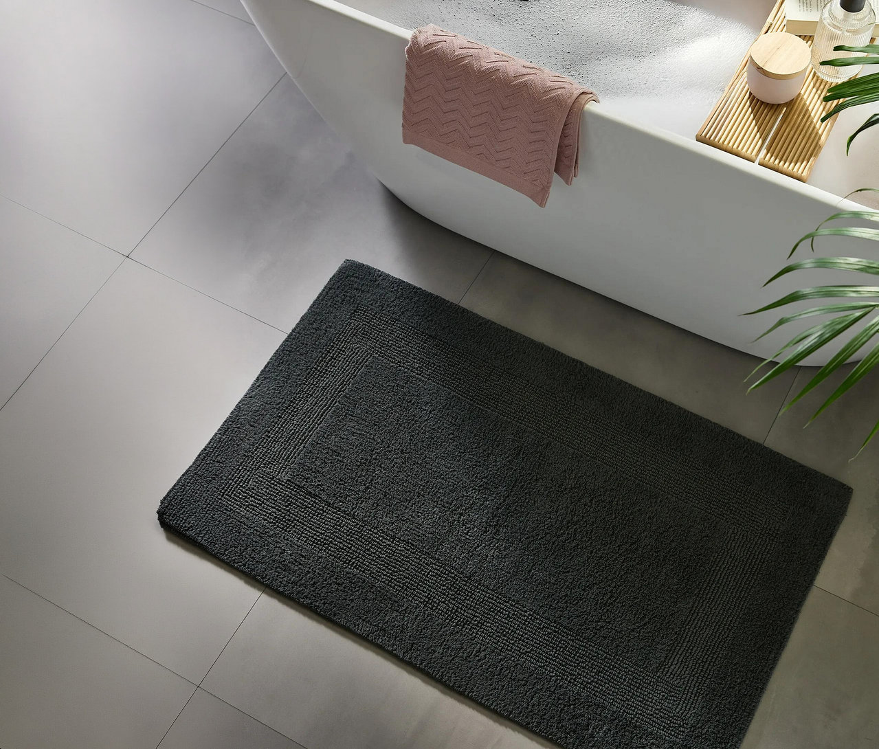 Dunkelgraue Badematte auf hellgrauen Fliesen vor einer weißen Badewanne mit rosa Handtuch, Badeschaum, einem Holzbadebrett und einer Pflanze.