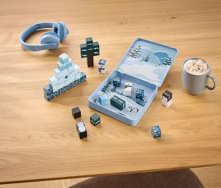 Hellblaue Kopfhörer, ein offenes Winter-Spielset mit Bausteinen, die ein Iglu und einen Baum bilden, sowie eine Tasse Kakao mit Marshmallows auf einem Holztisch.