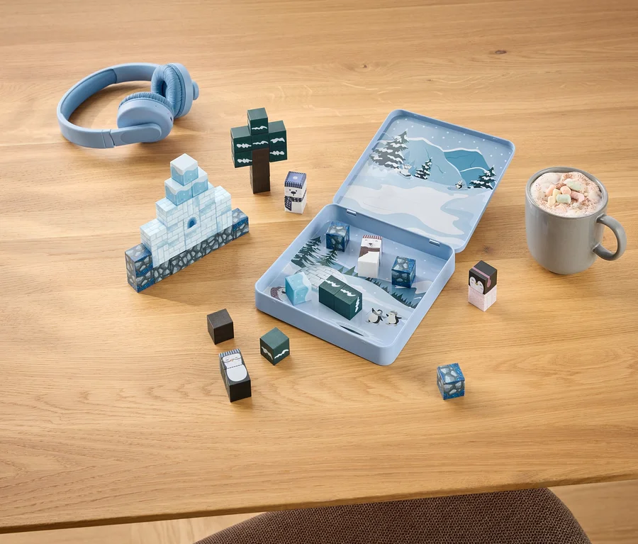 Hellblaue Kopfhörer, ein offenes Winter-Spielset mit Bausteinen, die ein Iglu und einen Baum bilden, sowie eine Tasse Kakao mit Marshmallows auf einem Holztisch.