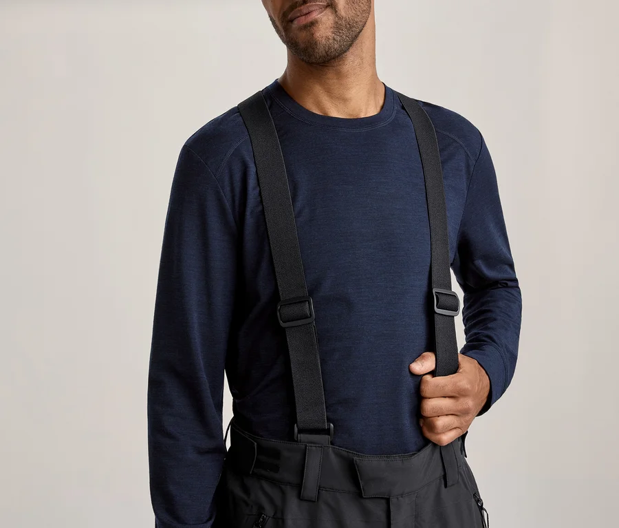 Mann trägt ein dunkelblaues Langarmshirt und eine schwarze Hose mit Hosenträgern, wobei er einen Hosenträger mit der rechten Hand hält.