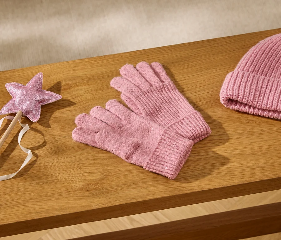 Rosa Glitzerstern-Zauberstab mit Bändern, rosa Strickhandschuhe und eine rosa Strickmütze auf einer Holzoberfläche.