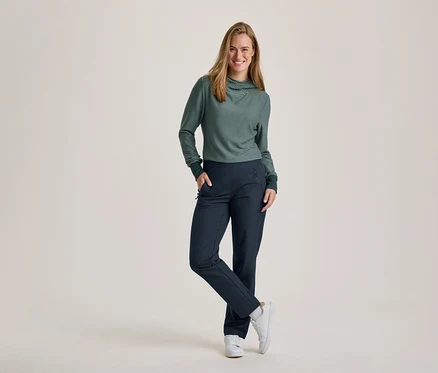 Eine lächelnde Frau mit langen Haaren steht mit einer Hand in der Tasche und einem gekreuzten Bein. Sie trägt ein grün-weiß gestreiftes Langarmshirt mit Rollkragen und dunkelblaue Hosen sowie weiße Turnschuhe.