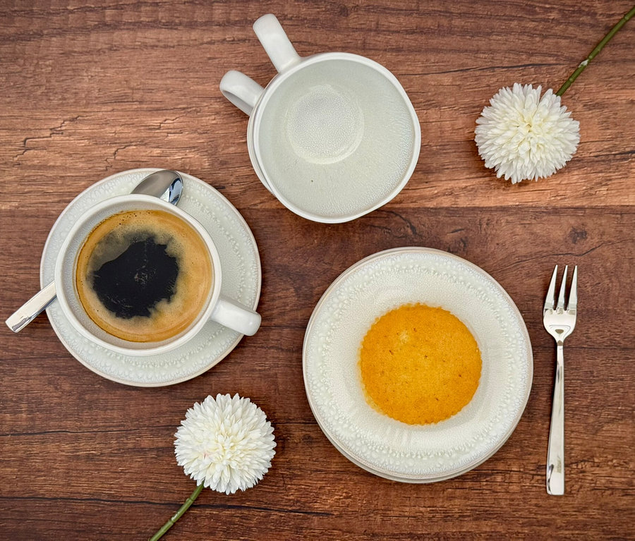 Flat Lay: Helles Mäser Kaffeetassen-Set »Frozen« mit schwarzem Kaffee, leerer Tasse, Teller mit gelbem Muffin und Gabel, dazu zwei weiße Deko-Blumen auf Holztisch.