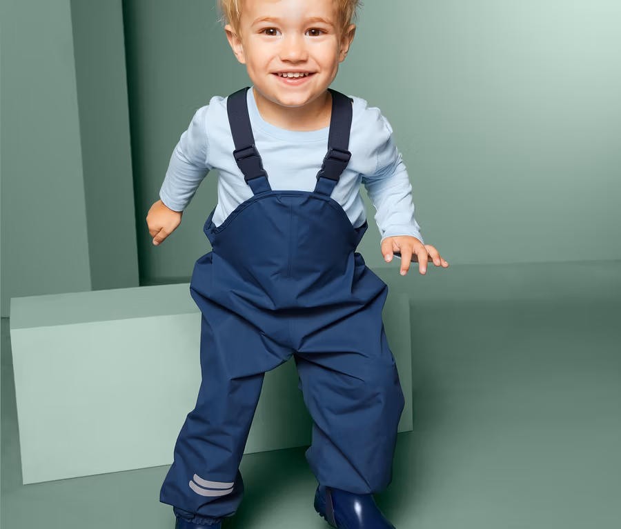 Ein lächelndes Kleinkind steht und trägt eine blaue Regenhose mit Latz und blaue Gummistiefel.
