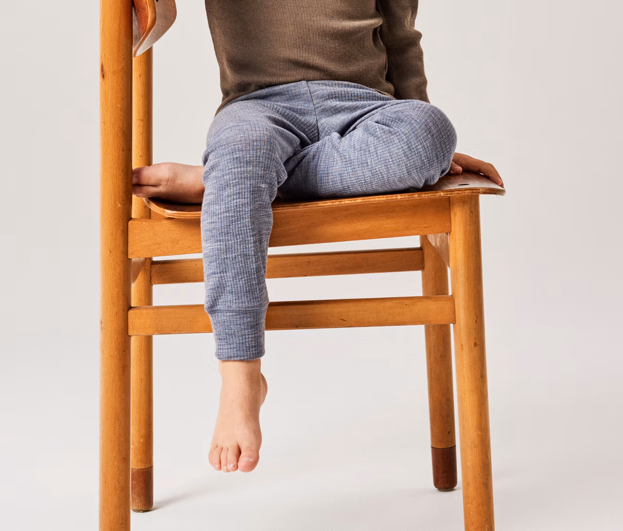 Kind sitzt auf einem Holzstuhl, trägt ein braunes Langarmshirt und eine blaue gemusterte Hose, ein Fuß baumelt.