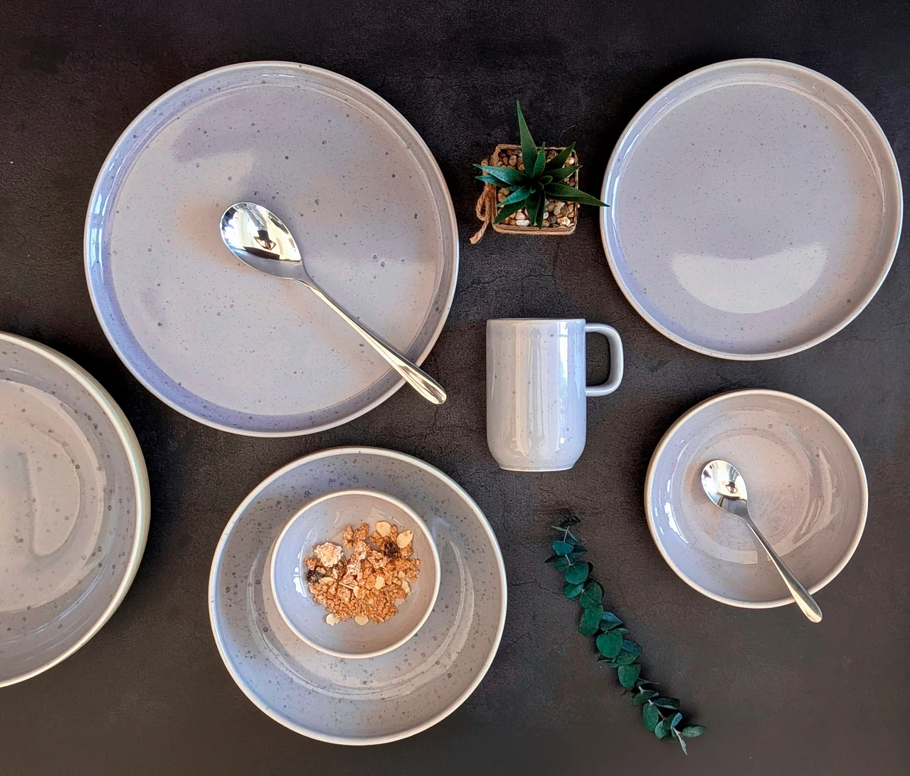 Helles, grau gesprenkeltes Mäser Tafelservice »Candy«, 8-tlg, auf dunklem Untergrund. Es umfasst Teller, Schüsseln, einen Becher und Löffel. Eine Schüssel enthält Müsli, daneben eine kleine Pflanze und Eukalyptuszweige.