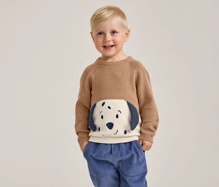 Ein lächelnder kleiner Junge mit blondem Haar steht mit den Händen in den Taschen und trägt einen braunen Pullover mit einem weißen und blauen Hundegesicht auf der Vorderseite sowie eine blaue Cordhose.