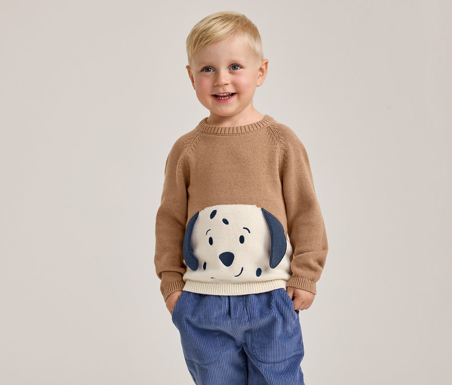 Ein lächelnder kleiner Junge mit blondem Haar steht mit den Händen in den Taschen und trägt einen braunen Pullover mit einem weißen und blauen Hundegesicht auf der Vorderseite sowie eine blaue Cordhose.