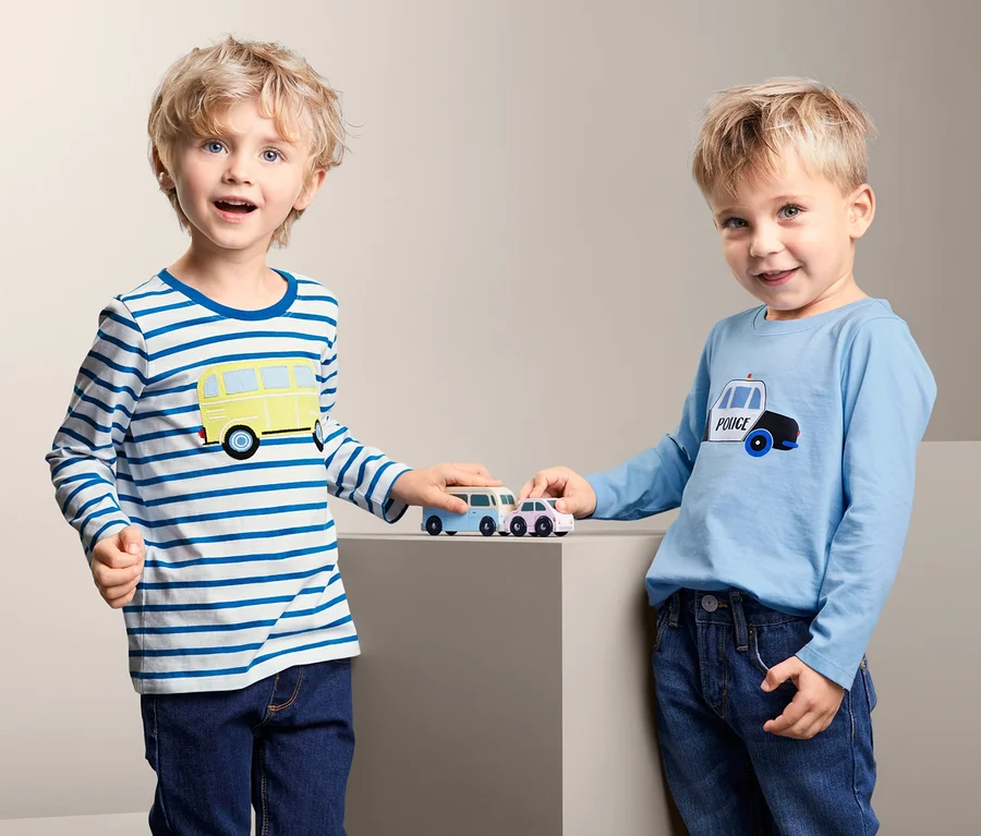 Zwei Jungen spielen mit Spielzeugautos. Einer trägt ein 2 Langarmshirts mit Busaufdruck, der andere ein Langarmshirt mit Polizeiauto.