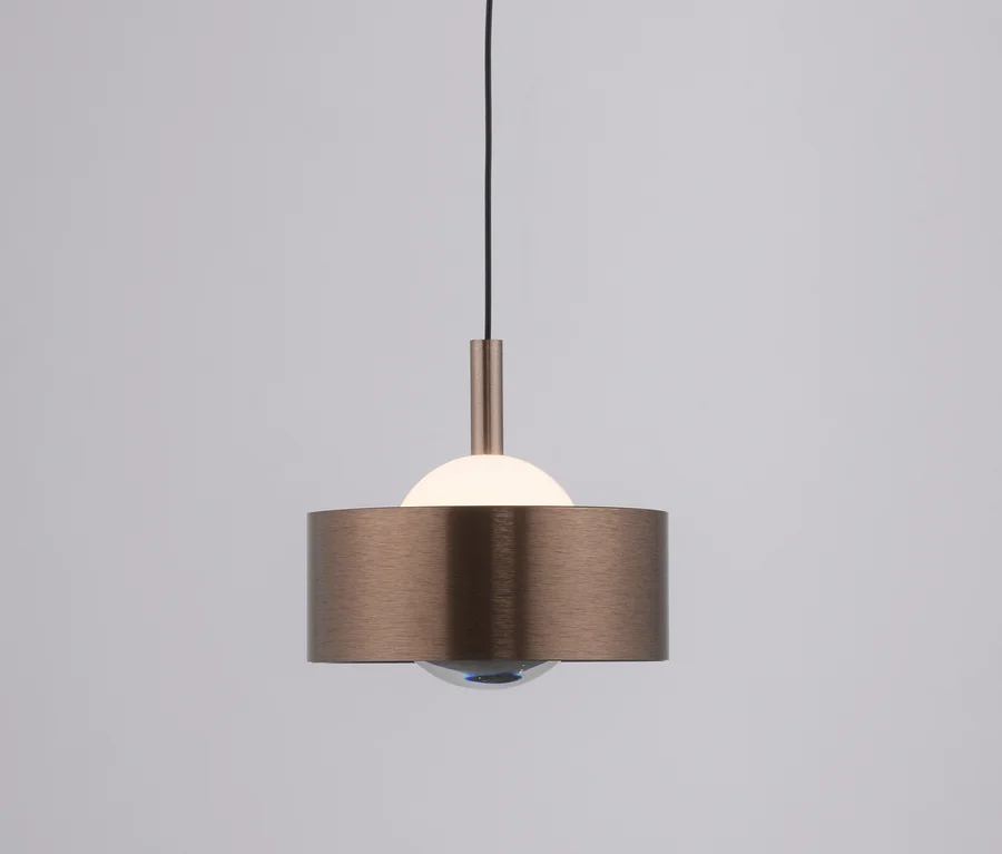 Eine Pendelleuchte mit zylindrischem Schirm aus gebürsteter Bronze, einer leuchtenden weißen Halbkugel oben und einer klaren Kugel unten, hängt an einem schwarzen Kabel.
