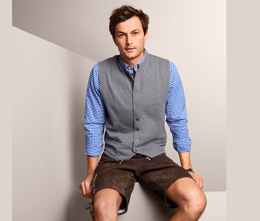 Mann sitzt und trägt eine Sweatweste, ein Hemd und eine Lederhose.