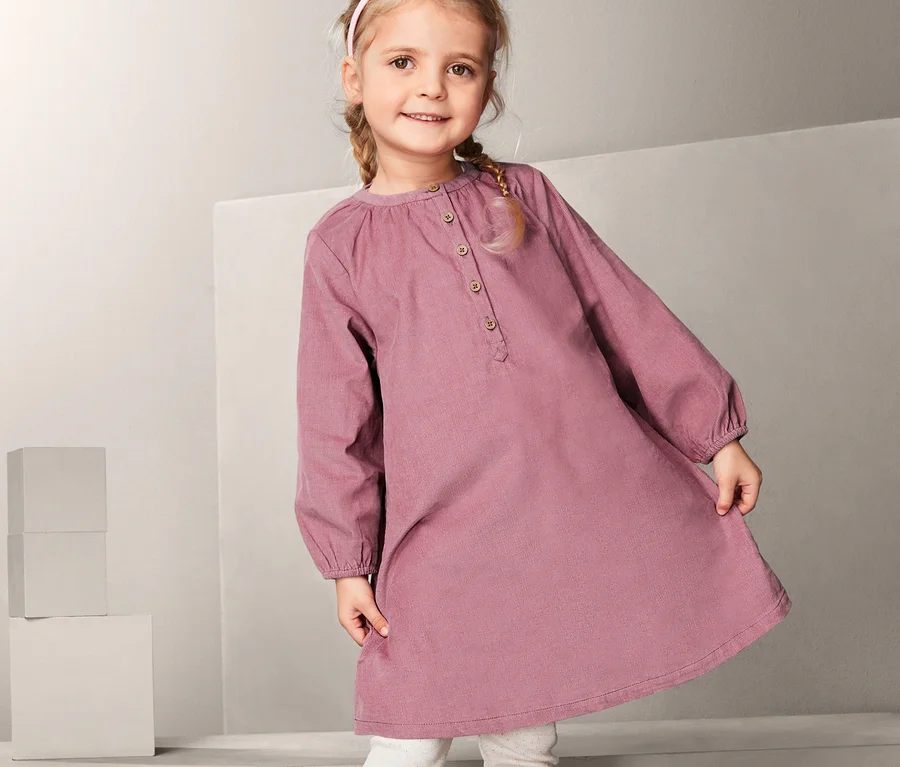 Mädchen steht auf einem weißen Würfel und trägt ein Cordkleid, weiße Leggings und braune Schuhe.