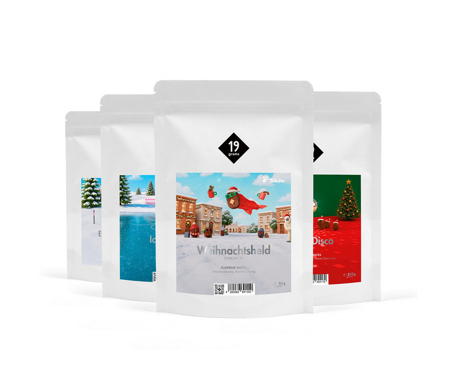 Vier weiße Kaffeepackungen stehen schräg hintereinander. Die vorderste Packung zeigt einen fliegenden Weihnachtshelden über einer verschneiten Stadt und ist mit „Weihnachtsheld“ beschriftet. Rechts daneben ist eine Packung mit „Disco“ und einem Weihnachtsbaum zu sehen. Links sind zwei weitere Packungen mit Wintermotiven.